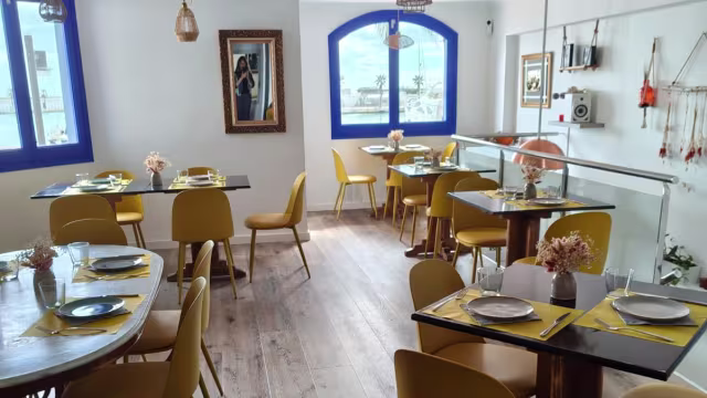 Interior de restaurante moderno y luminoso, con paredes claras, grandes ventanales y mesas decoradas con servilletas amarillas. Sillas elegantes amarillas y elementos decorativos como espejos y obras de arte.