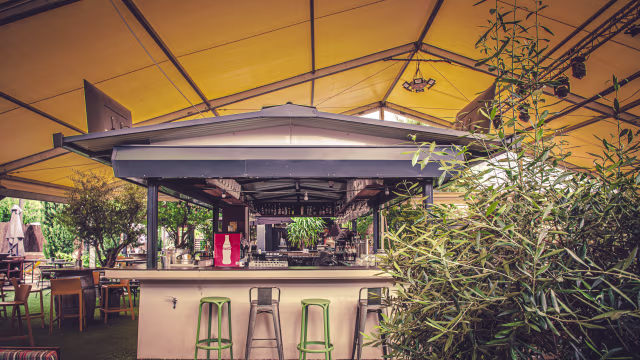 Espace de restauration extérieur spacieux avec un grand auvent jaune, un bar élégant et des plantes vertes luxuriantes, créant une atmosphère accueillante et détendue.