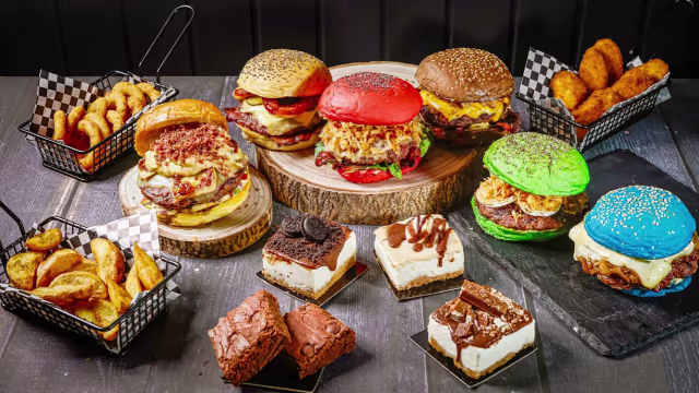 Variedade de hambúrgueres gourmet com pães coloridos, recheados com queijo derretido, alface fresca e diversos toppings, acompanhados de batatas fritas crocantes. Inclui também quadrados de sobremesa, como brownies ricos e camadas cremosas com cobertura de chocolate e migalhas de bolacha.
