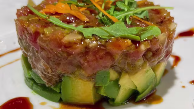 Tartar de atún finamente picado sobre una cama de aguacate cremoso, decorado con hierbas frescas y hebras de naranja, acompañado de una reducción de salsa de soja o sésamo.