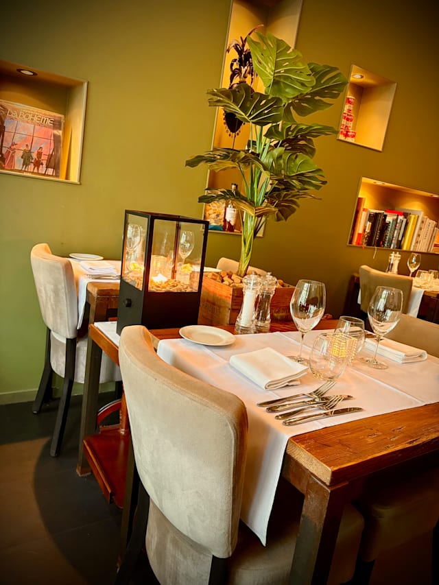 Een modern restaurantinterieur met houten tafels, witte tafelkleden en netjes gerangschikt bestek. De groene muren en zachte verlichting creëren een warme sfeer, terwijl een hoge kamerplant een vleugje groen toevoegt. Planken met boeken en decoratieve items verfraaien het geheel, wat wijst op een comfortabele en uitnodigende eetervaring.