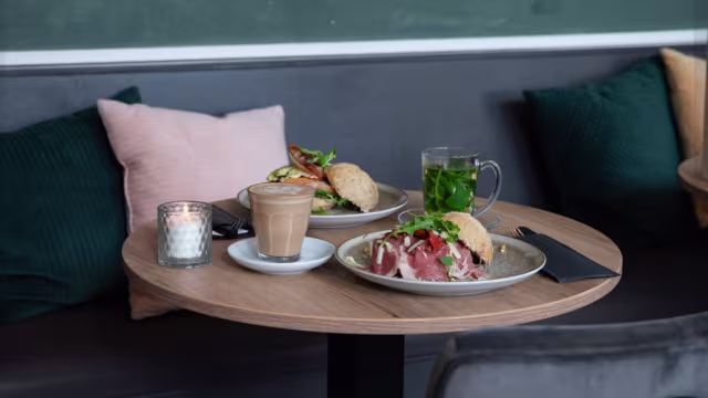 Een gezellige tafelsetting met een frisse salade van gemengde groenten, komkommer en cherrytomaten, en een stevig gerecht met dunne plakken vlees, mogelijk prosciutto, gegarneerd met verse kruiden en een zacht broodje. Vergezeld door een groen drankje en een kop koffie of thee.
