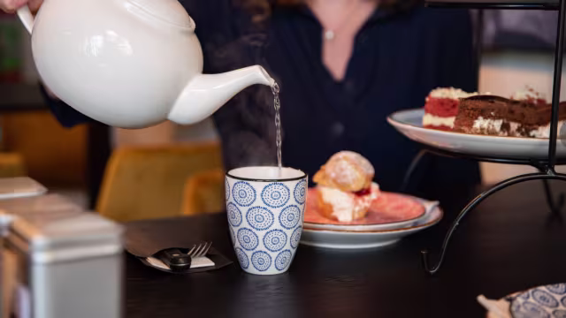 Een dampende kop thee wordt ingeschonken uit een witte theepot in een blauw-witte kop. Op de tafel staan delicate gebakjes, waaronder een scone met room en jam, en een selectie van taarten met lagen van chocolade en room. De sfeer straalt een gezellige afternoon tea uit, die uitnodigt tot ontspanning en genot.