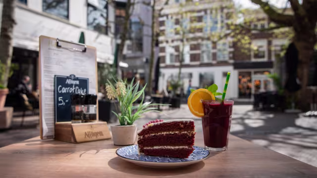 Een plak worteltaart, gelaagd met roomkaasglazuur, met een vochtige textuur en zichtbare wortelreepjes en specerijen zoals kaneel en nootmuskaat. Vergezeld van een verfrissende fruitdrank, gegarneerd met een schijfje sinaasappel.