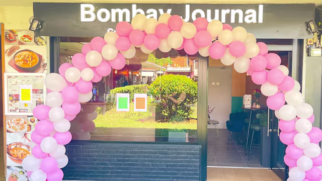 Entrée accueillante avec un arc décoratif en ballons roses et blancs, portant le nom 'Bombay Journal', accompagnée d'un tableau de menu et d'une ambiance lumineuse visible à travers de grandes fenêtres.