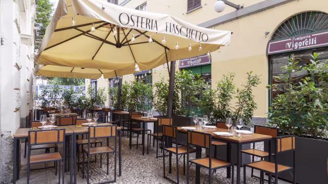 Area esterna di un ristorante con tavoli e sedie in legno sotto un grande tendone, decorata con piante in vaso e un'atmosfera accogliente, dedicata alla cucina italiana.