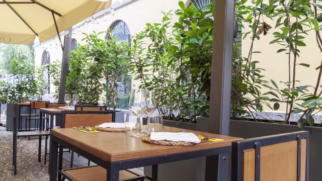 Area pranzo all'aperto con tavoli in legno sotto un grande ombrello, circondati da vegetazione lussureggiante, creando un'atmosfera invitante e rilassata.