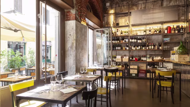 Interni moderni e rustici con pareti in mattoni a vista e ampie finestre, tavoli in legno e sedie colorate, creando un'atmosfera accogliente e informale.