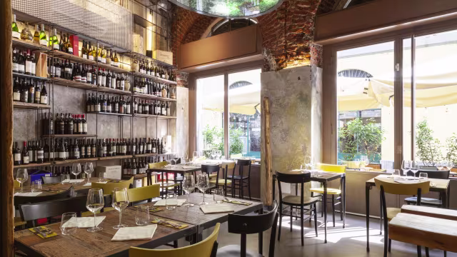 Interno accogliente di un ristorante con atmosfera calda, pareti decorate con scaffali di bottiglie di vino, ampie finestre che illuminano tavoli e sedie in legno.