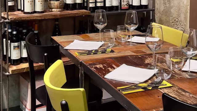 Impostazione rustica con tavolo in legno, sedie nere moderne e alcune sedie gialle, circondato da una selezione di bottiglie di vino.