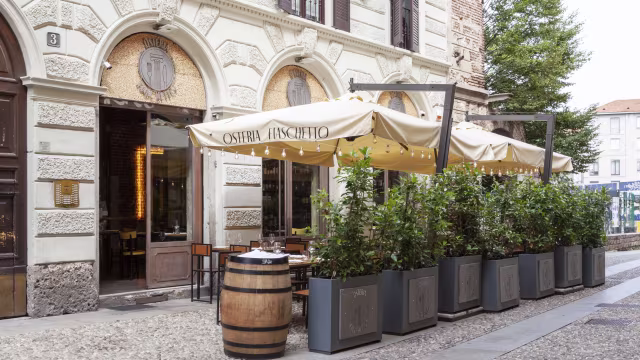 Ristorante con un'atmosfera accogliente, ingresso con porte in vetro, tavoli sotto un'ombrellone beige e circondato da verde lussureggiante.