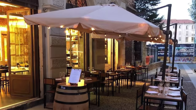 Area esterna accogliente con tavoli in legno sotto ombrelloni, illuminazione soffusa e atmosfera calda per un'esperienza di ristorazione all'aperto.