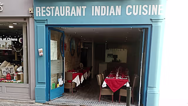 Façade bleu vif avec une enseigne 'RESTAURANT INDIAN CUISINE', entrée ouverte sur un intérieur chaleureux avec des tables bien disposées et des nappes rouges.