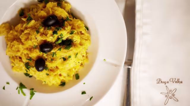 Um montículo de mandioca desfiada, dourada e fofa, temperada com especiarias, guarnecido com ervas verdes picadas e azeitonas pretas, que acrescentam um toque salgado.