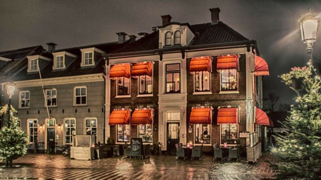 Charmante restaurantgevel bij nacht met een twee verdiepingen tellend gebouw in klassieke stijl, grote ramen, feloranje luifels en een uitnodigende buitenzitplaats.