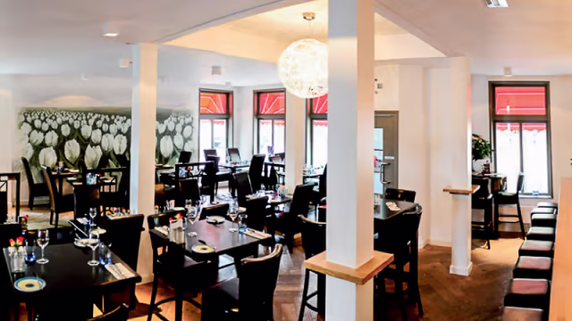 Het interieur van het restaurant heeft een moderne en ruime indeling met houten vloeren en een mix van donkere houten tafels en stoelen. De muren zijn versierd met een groot muurschildering van witte tulpen, wat een vleugje elegantie toevoegt aan de sfeer. Natuurlijk licht valt binnen via grote ramen, wat een warme ambiance creëert. De zitgelegenheid omvat zowel tafels als een lange bank, wat verschillende eetervaringen mogelijk maakt. Het ontwerp combineert hedendaagse elementen met een gastvrije uitstraling, waardoor het geschikt is voor zowel informele als speciale gelegenheden.