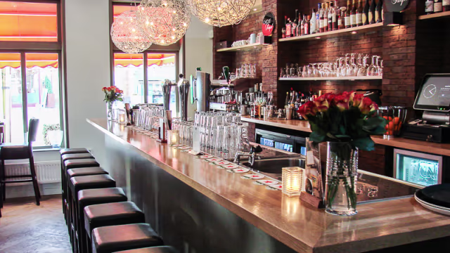 Stijlvolle bar met een gepolijst houten aanrecht, verlichte flessen achter de bar en moderne zwarte krukken, aangevuld met verse bloemen voor een uitnodigende sfeer.