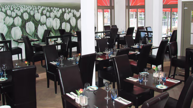 Het interieur van het restaurant heeft een ruime indeling met donkere houten tafels en elegante zwarte stoelen. De muren zijn versierd met een groot muurschildering van witte tulpen, wat een frisse en uitnodigende sfeer creëert. Natuurlijk licht stroomt binnen via grote ramen, aangevuld met helderrode zonneschermen aan de buitenkant. Elk tafel is netjes gedekt met bestek en glazen, wat de elegante maar informele eetervaring versterkt. De indeling suggereert een comfortabele setting voor zowel intieme maaltijden als grotere bijeenkomsten.