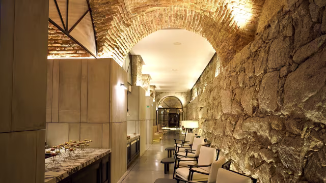 Interior de restaurante elegante com um corredor arqueado, paredes de pedra suave e acabamentos modernos, iluminação suave que destaca as características arquitetónicas e uma área de estar com cadeiras minimalistas.