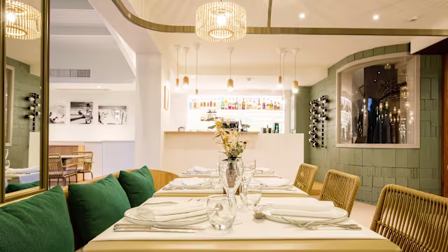 Intérieur de restaurant moderne avec une salle à manger bien éclairée, des tables élégamment dressées, des coussins verts et un bar chic, créant une atmosphère chaleureuse et accueillante.