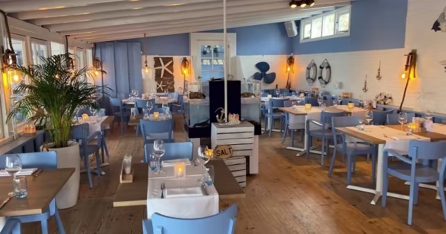 Het interieur van het restaurant heeft een kustthema met lichtblauwe muren en houten accenten, wat een ontspannen sfeer creëert. De ruimte is goed verlicht met warme hanglampen en is versierd met nautische decoraties zoals touwen en ankers, wat bijdraagt aan de zee-atmosfeer.