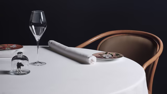 Tavolo da pranzo elegantemente apparecchiato con tovaglia bianca, bicchiere di vino, tovagliolo arrotolato e un piccolo campana di vetro che copre un delicato piatto.