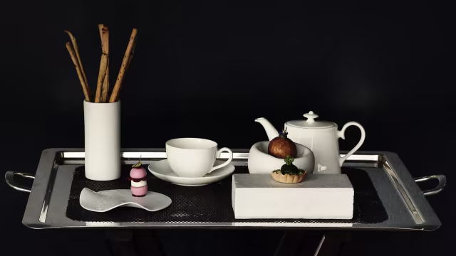 Vassoio elegante con teiera bianca, tazza abbinata, pasticcino al cioccolato, dessert a strati rosa e bianco, e grissini croccanti.