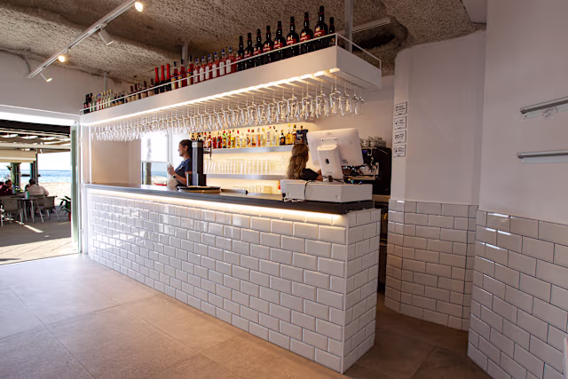 Bar moderno con mostrador de azulejos blancos, iluminación brillante y estanterías organizadas con botellas.