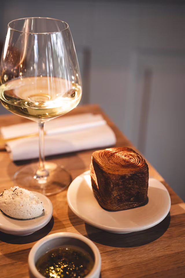 Mooi gepresenteerd gerecht met een vierkant stuk versgebakken brood met een spiraalpatroon en een goudbruine korst, geserveerd met romige boter met een snufje zout en een groene kruiden saus, waarschijnlijk pesto. Een glas witte wijn maakt de verfijnde eetervaring compleet.