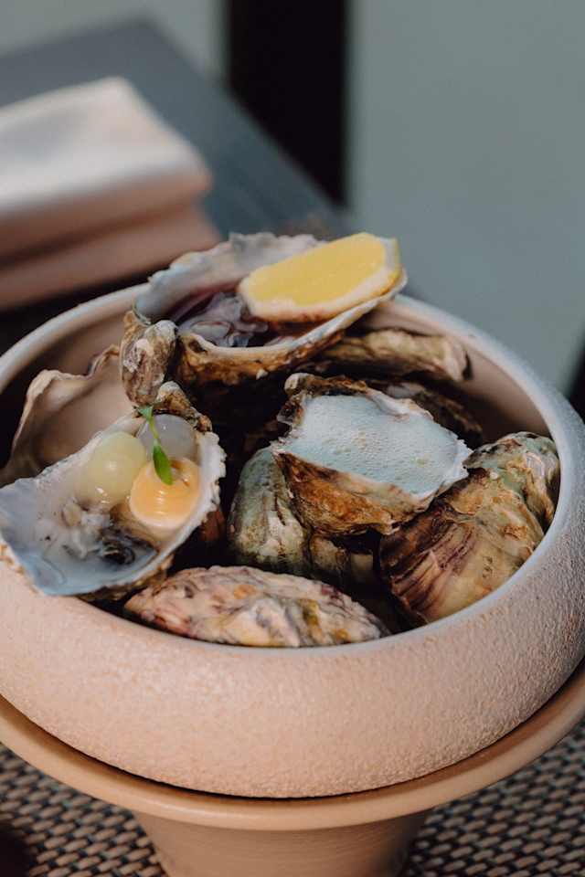 Een kom met verse oesters in hun natuurlijke schelpen, gedeeltelijk geopend en met glanzend, stevig vlees. Sommige oesters zijn gegarneerd met kleine toefjes saus, mogelijk op basis van citrus of azijn, en een takje groene kruiden voor extra frisheid. De presentatie benadrukt de versheid en kwaliteit van de zeevruchten.