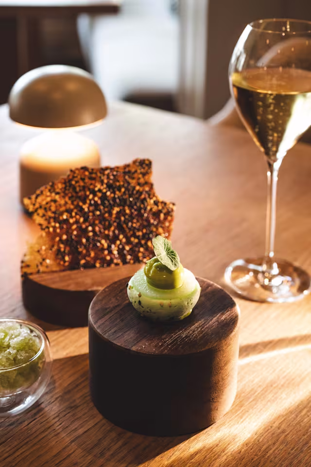 Een verfijnd gerecht met een kleine, ronde groene mousse, gegarneerd met een groene olijf en een muntblaadje. Geserveerd met een klein kommetje fijngehakte groene garnering en een stuk geroosterde knapperige textuur op de achtergrond.