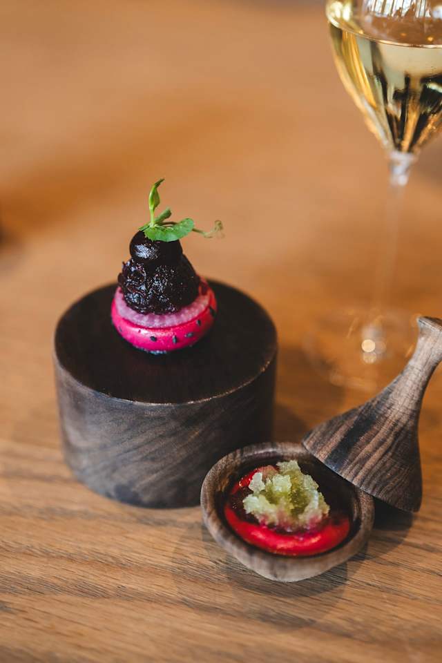 Een visueel opvallend gerecht met een kleine, ronde hoop van donkere, rijke ingrediënten, mogelijk een mousse of purée, gegarneerd met een delicate groene kruiden. Daaronder bevindt zich een levendige roze laag, waarschijnlijk gemaakt van een fruit of groente. Bij het hoofdgerecht hoort een klein houten schaaltje met een lichte, textuurvolle condiment, dat lijkt op een relish of saus in een heldere rode kleur. De presentatie is elegant en artistiek, wat wijst op een creatieve culinaire benadering.