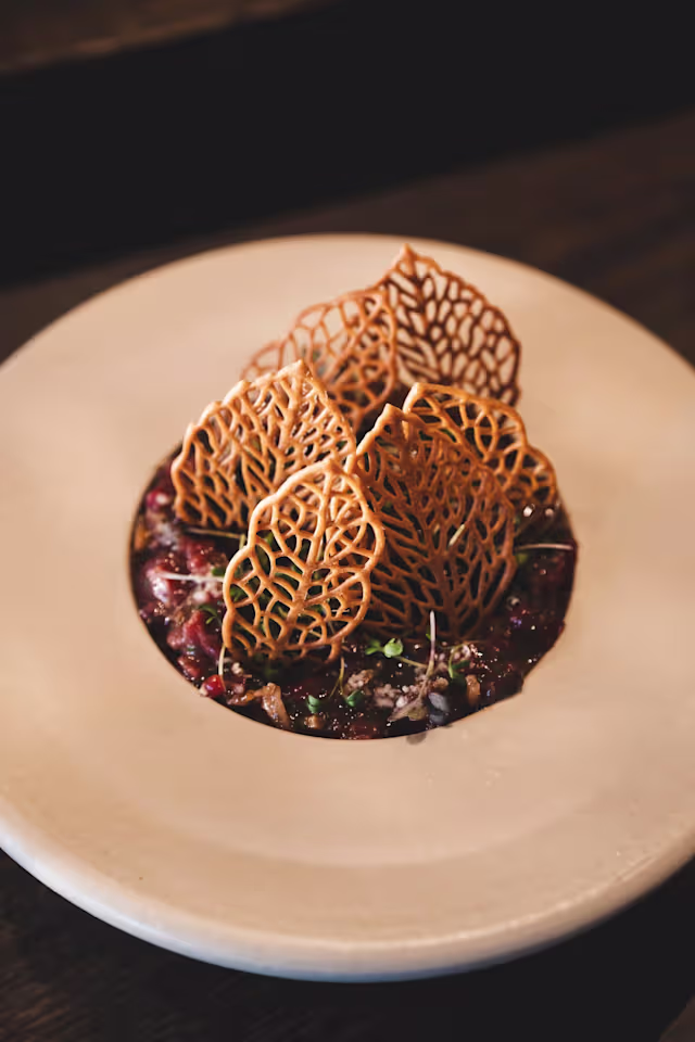 Mooi gepresenteerd gerecht met fijngehakt rauw vlees, waarschijnlijk rundvlees, geserveerd met een rijke saus in diepe rode en bruine tinten. Gegarneerd met delicate, knapperige versieringen die op bladeren lijken, en bestrooid met kleine kruiden, wat de frisheid en visuele aantrekkingskracht van het gerecht vergroot.