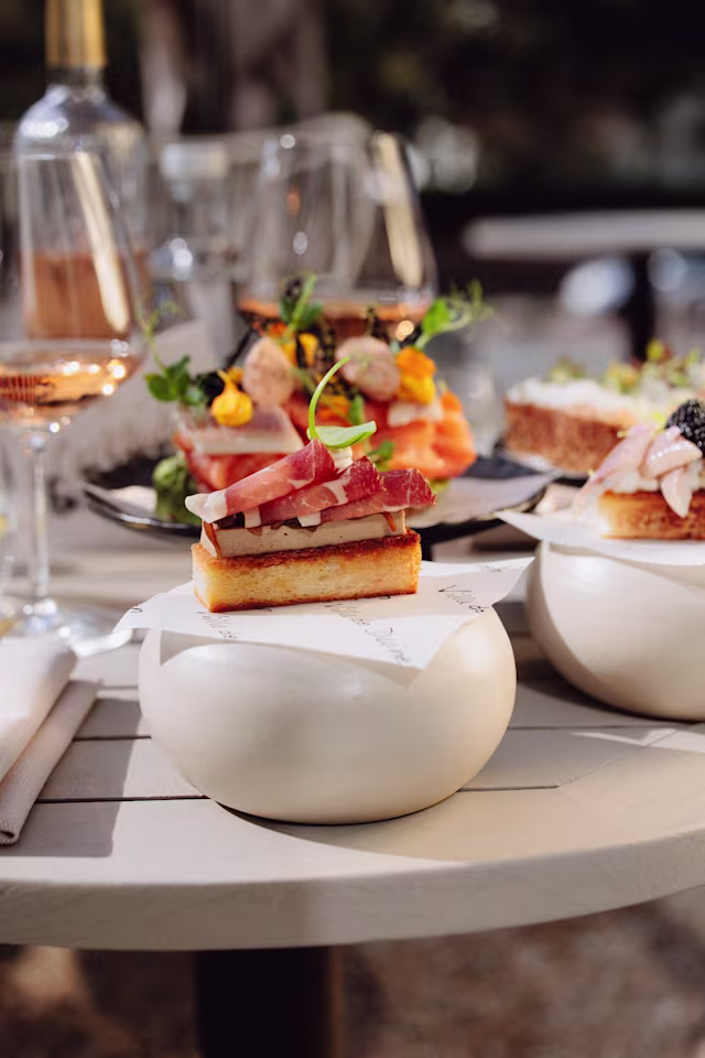 Een prachtig gepresenteerde selectie van gourmetcanapés, waaronder een rechthoekig stuk brood met een romige spread, dunne plakjes vlees en een takje kruiden. Achtergrond toont diverse canapés met zeevruchten en kleurrijke garneringen.