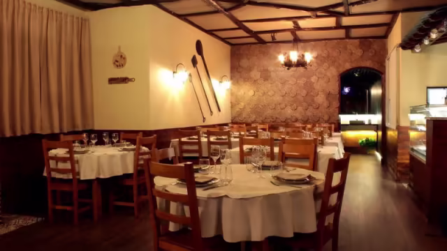 Interior do restaurante com uma atmosfera acolhedora, mesas e cadeiras de madeira, paredes com design texturizado e utensílios de madeira como decoração, iluminação suave e janelas grandes que permitem a entrada de luz natural.