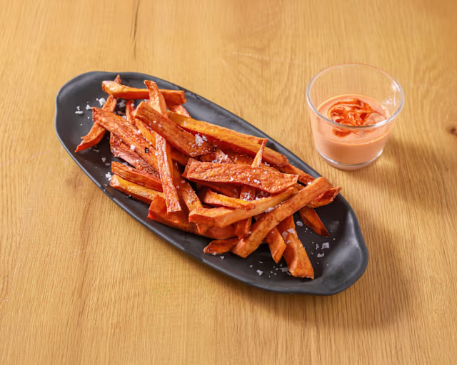 Porciones generosas de papas fritas de batata doradas y crujientes, ligeramente espolvoreadas con sal marina y acompañadas de una salsa cremosa con un toque de naranja.