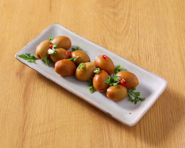 Plato de aceitunas marinadas en tonos marrones y verdes, decoradas con trozos de chile rojo y perejil fresco.