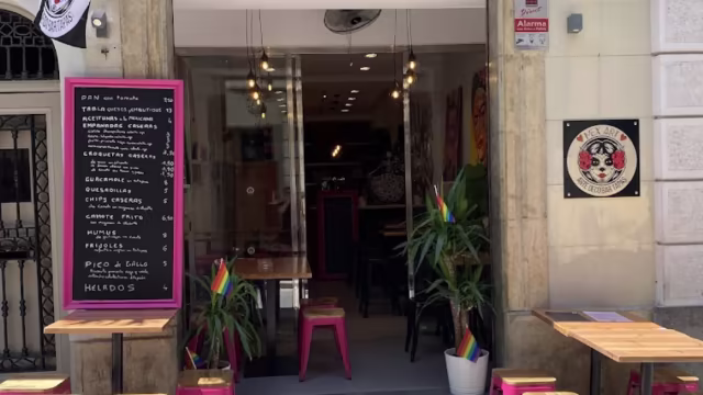 Exterior de un restaurante vibrante con un ambiente acogedor, menú de pizarra que destaca platos mexicanos como tacos, burritos y nachos, y mesas de madera con taburetes rosas.