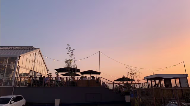 Het beeld toont een restaurantsetting tijdens zonsondergang, met een moderne buiten eetruimte. De structuur heeft een glazen gedeelte voor een panoramisch uitzicht. Verschillende tafels staan onder grote parasols voor schaduw, terwijl sfeerverlichting boven de tafels een warme ambiance creëert. De lucht is gekleurd in zachte tinten van oranje en paars, wat bijdraagt aan de charme van de scène.