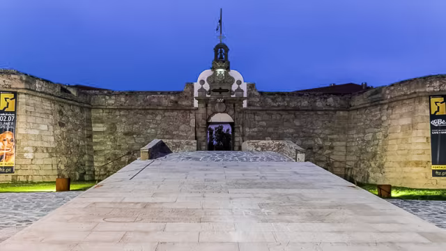 Estrutura histórica de pedra, possivelmente um forte ou castelo, com um arco central e paredes altas de pedra desgastada, apresentando uma mistura de tons claros e escuros. O caminho de pedras planas leva à entrada, enquanto o céu ao crepúsculo realça os detalhes arquitetónicos.