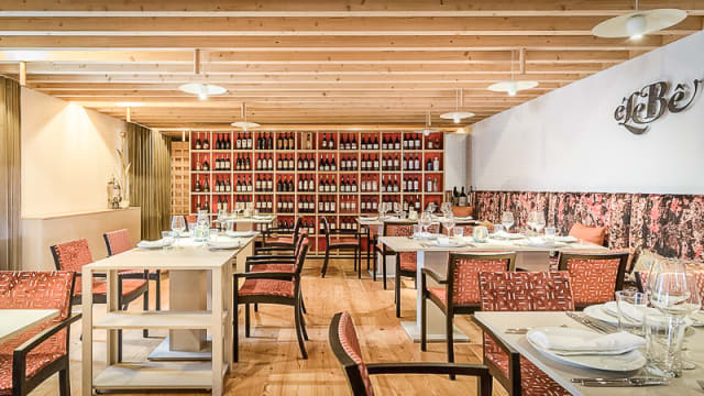 Interior moderno com vigas de madeira e palete de cores quentes, mesas de madeira clara e cadeiras estofadas em tecido rico. Uma parede destaca uma coleção de garrafas de vinho dispostas em grelha, criando um ambiente sofisticado e acolhedor.