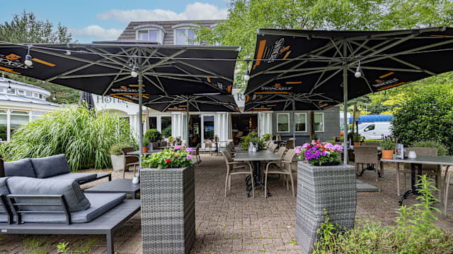 Buiten eetgedeelte van een restaurant met tafels onder grote, donkere parasols, comfortabele lounge stoelen en omringd door weelderig groen, met bloempotten vol kleurrijke bloemen voor een uitnodigende en ontspannen sfeer.
