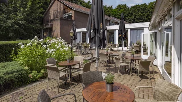 Buiten eetgedeelte van een restaurant met ronde houten tafels en rieten stoelen, omringd door weelderig groen en bloeiende planten, met grote parasols voor schaduw.