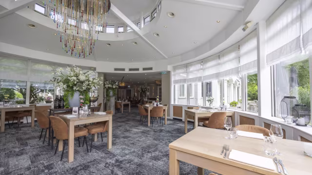 Het interieur van het restaurant heeft een ruime en moderne uitstraling met grote ramen die natuurlijk licht binnenlaten. De decoratie omvat elegante bloemstukken die frisheid toevoegen. De tafels zijn gedekt met eenvoudige maar stijlvolle serviezen, en de zitplaatsen bestaan uit comfortabele stoelen. De algehele sfeer is uitnodigend en verfijnd, perfect voor een ontspannen eetervaring.