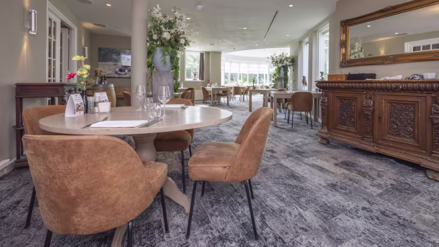 Een verfijnd restaurantinterieur met elegante houten meubels en luxe, tan kleurige stoelen. Het is goed verlicht met natuurlijk licht dat door grote ramen binnenstroomt, wat een warme en uitnodigende sfeer creëert. Verse bloemstukken sieren de tafels en voegen kleur en frisheid toe. De decoratie omvat smaakvolle kunstwerken aan de muren en een prachtig vervaardigde houten sidetable, wat de algehele verfijnde esthetiek versterkt.