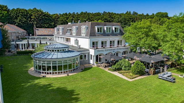 Een goed onderhouden restaurantgevel met een groot, elegant gebouw en een moderne glazen structuur. De omgeving is weelderig met groen, waaronder bomen en verzorgde gazons. Buiten zitplaatsen zijn netjes gerangschikt op een terras, met tafels en parasols voor schaduw. De algehele setting suggereert een serene en uitnodigende eetervaring in een schilderachtige omgeving.
