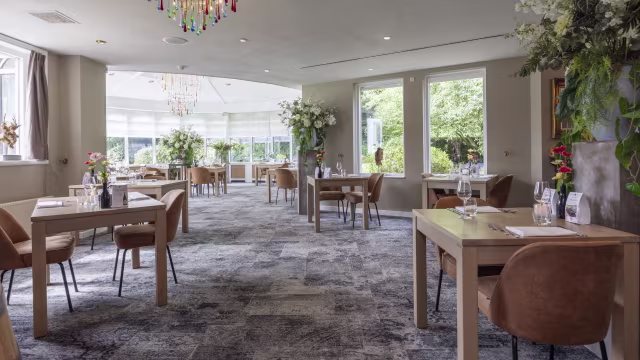 Ruimtelijk restaurantinterieur met moderne houten tafels en comfortabele bruine stoelen, verlicht door een kleurrijke kroonluchter. Grote ramen zorgen voor natuurlijk licht, terwijl elegante bloemstukken en groen de uitnodigende sfeer versterken. Het tapijt heeft een subtiel patroon dat bijdraagt aan het eigentijdse ontwerp.