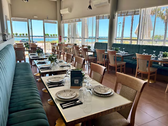 Interior de restaurante luminoso con grandes ventanales, banqueta verde y mesas de madera, creando un ambiente acogedor.
