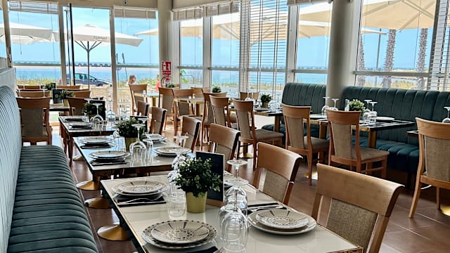 Interior espacioso con grandes ventanales que permiten la entrada de luz natural, decorado con plantas en macetas y una mezcla de sillas de madera y un banquete cómodo, creando un ambiente acogedor.