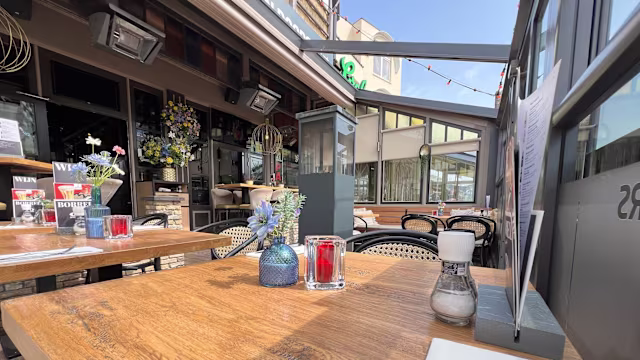Een charmante buiten eetruimte met houten tafels en decoratieve items zoals een blauwe glazen vaas met verse bloemen en een rode kaarsenhouder. De klassieke bistrostoelen met gevlochten zittingen zorgen voor een gezellige sfeer, verlicht door bovenverlichting, met grote ramen en een moderne gevel op de achtergrond.